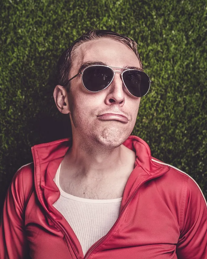 Hombre con gafas de sol de aviador y chándal rojo, tumbado en el césped con gesto de suficiencia
