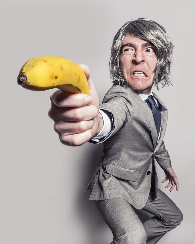 Hombre con traje gris y gesto de furia que apunta a la cámara con un plátano como si fuera una pistola