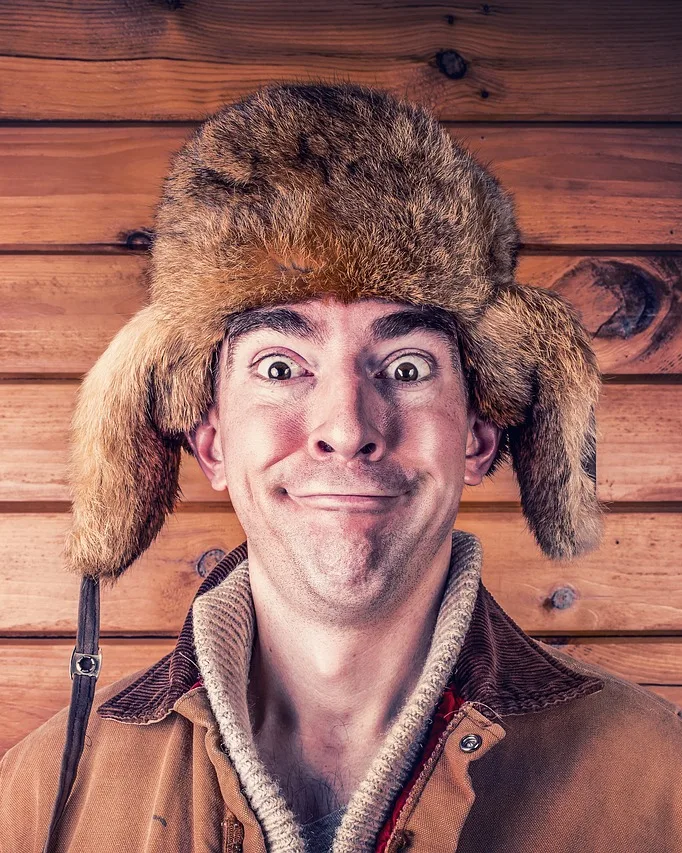 Hombre con un gorro de piel con orejeras y una sonrisa exagerada, delante de una pared de madera