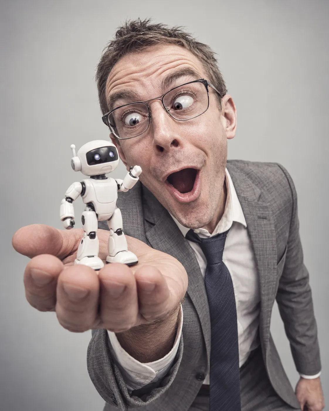 Hombre con traje gris y gafas que mira bizco y con la boca abierta a un pequeño robot blanco que sostiene en la palma de la mano
